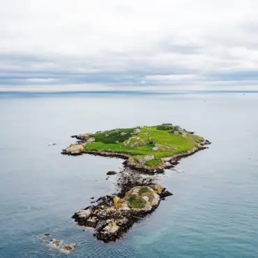 A small, rocky island with green grass is surrounded by calm blue water under a cloudy sky. The island is isolated, with no visible structures except some low ruins blending into the landscape.