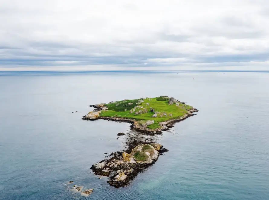 A small, rocky island with green grass is surrounded by calm blue water under a cloudy sky. The island is isolated, with no visible structures except some low ruins blending into the landscape.