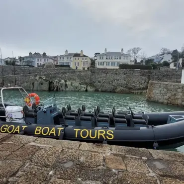 A black inflatable boat labeled GOAT BOAT TOURS is moored at a stone dock in a small harbor, with houses and trees visible on a hill in the background under a cloudy sky.