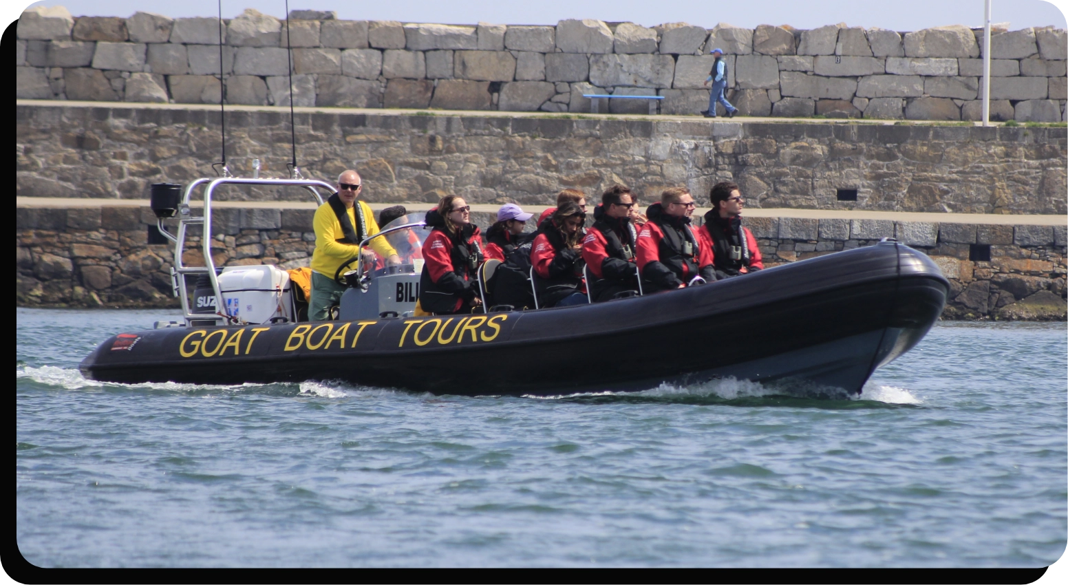 A group of people wearing life jackets are seated in a black inflatable boat labeled GOAT BOAT TOURS on the side. A person in a yellow jacket is steering the boat, which is moving through the water near a stone wall.