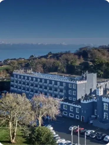 Aerial view of a large gray castle-like building with multiple windows and crenellated rooftops. The structure is surrounded by trees and a parking area with several cars. In the background, there is a vast body of water under a clear blue sky.