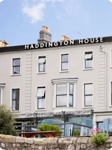 A three-story building with the sign HADDINGTON HOUSE in black letters at the top. The building is light gray with multiple windows, and a small seating area with plants is visible at the front. A seagull flies overhead.
