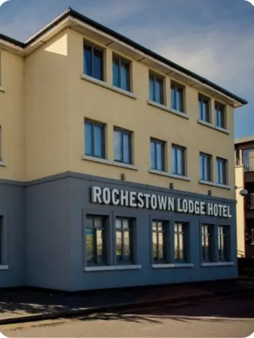 Street view of a three-story beige building with Rochestown Lodge Hotel signage. The building features multiple windows and a partial glimpse of another structure to the right. The sky is clear and blue.