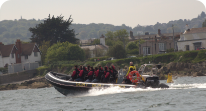 A black inflatable boat with several people in red life jackets speeds across the water near a shoreline with houses and trees in the background.