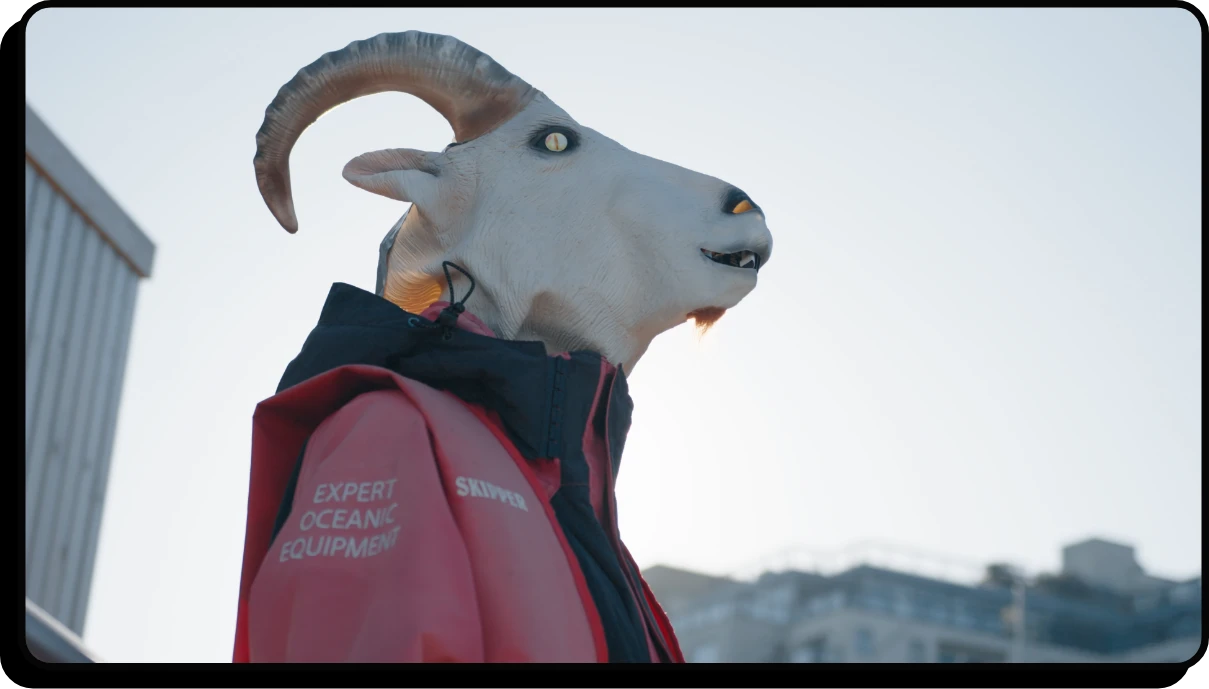 A person wearing a goat mask and a red jacket labeled EXPERT OCEANIC EQUIPMENT stands outdoors with a building in the background under a clear sky.