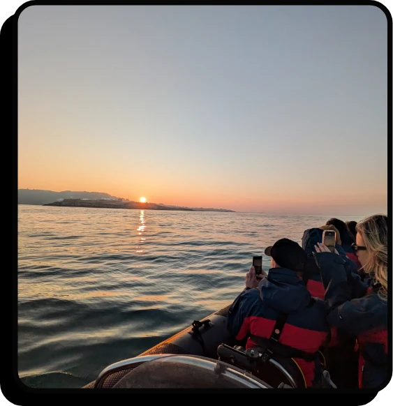 A group of people in life jackets sit in a boat on calm water, taking photos of the sun rising or setting over distant mountains under a clear sky.