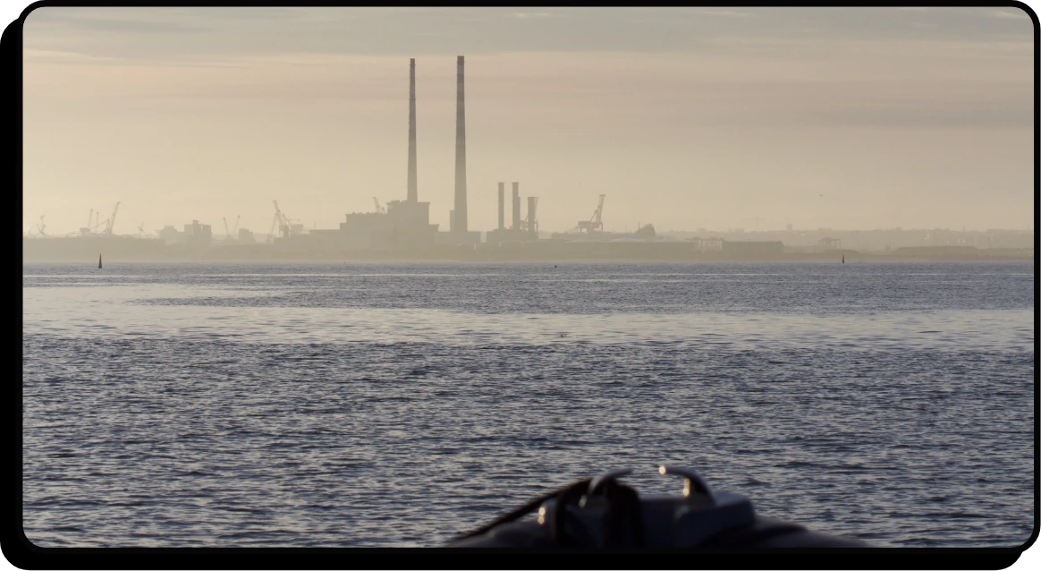 A calm body of water in the foreground with a blurred industrial landscape and tall smokestacks on the distant shoreline under a hazy sky.