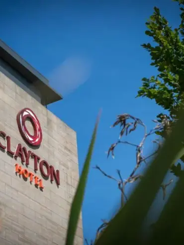 A modern hotel building with the sign Clayton Hotel in red letters, partially obscured by green leaves and branches, under a clear blue sky.