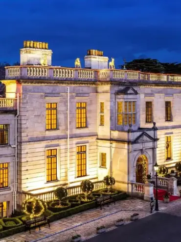 A grand, illuminated stone mansion at dusk, with large windows glowing warmly, manicured gardens, decorative lights, and a red carpet at the entrance, surrounded by trees and a clear evening sky.