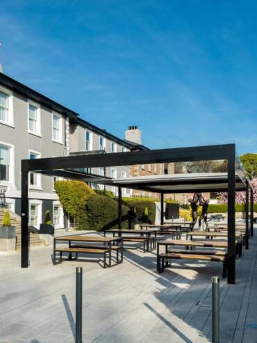 Outdoor seating area with wooden tables and benches under modern black pergolas, in front of a grey building with white trim, surrounded by hedges and trees on a sunny day.