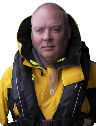 A person wearing a yellow waterproof jacket and a black life vest looks directly at the camera. The hood of the jacket is up, and the background is plain white.